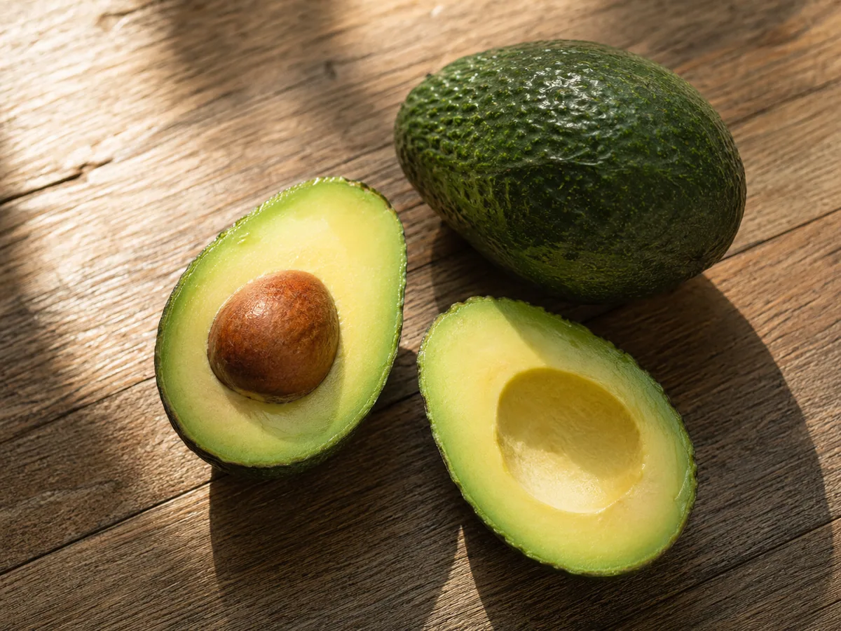 A ripe avocado cut in half beside a whole avocado on a sunlit wooden table.