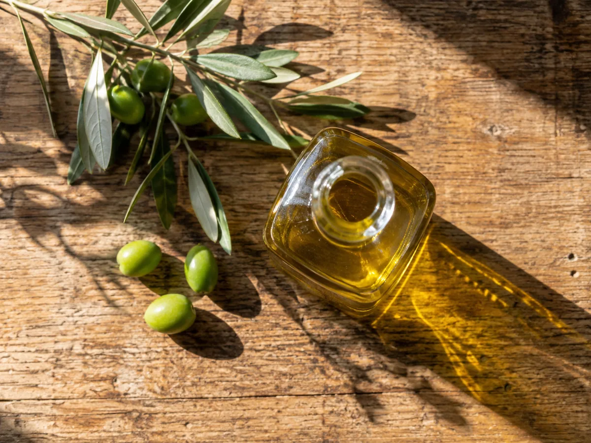 A glass bottle of extra-virgin olive oil on a rustic wooden table with an olive branch and fresh green olives.