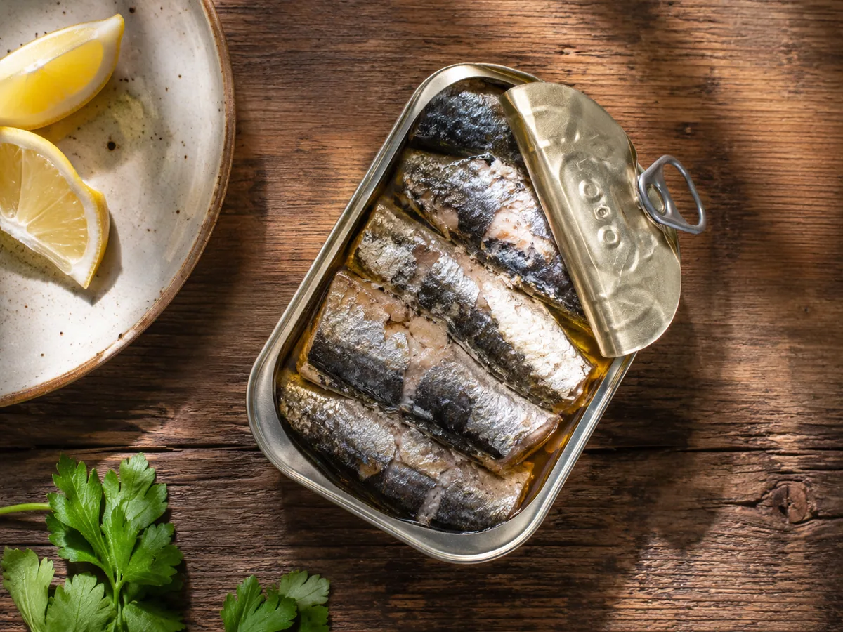 An open tin of sardines in oil on a wooden table with lemon wedges on a ceramic plate and fresh parsley.