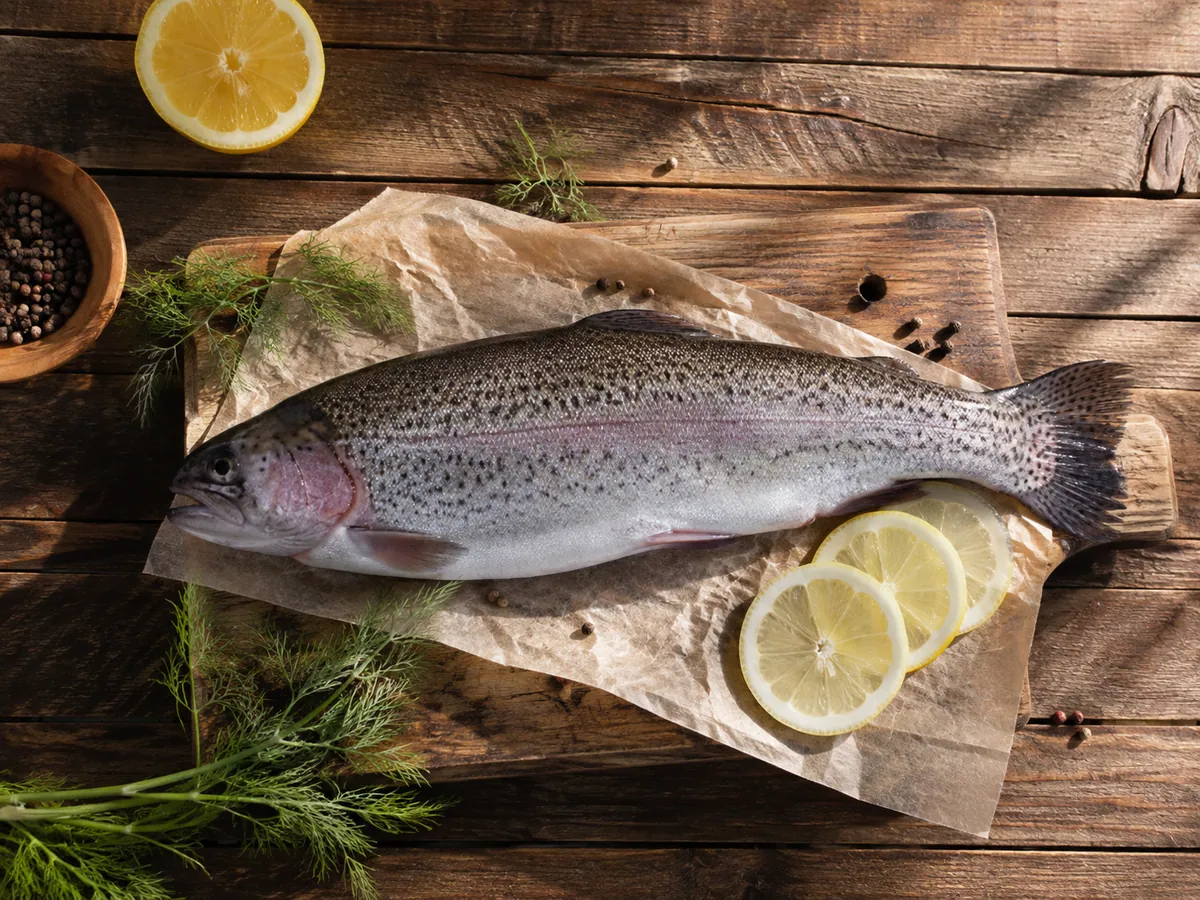 A whole cleaned trout on parchment paper over a wooden cutting board, with lemon slices, fresh dill, and a small bowl of peppercorns.